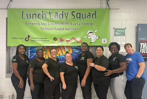 Lunch Ladies standing together in the cafeteria with a banner behind them that reads, Lunch Lady Squad